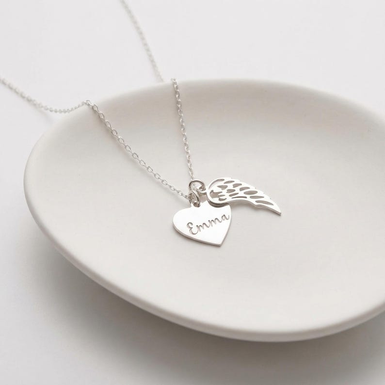 May include: A silver necklace with a heart pendant engraved with the name "Emma" and a wing charm. The necklace is displayed on a white, shallow dish. The chain and charms are bright and reflective.