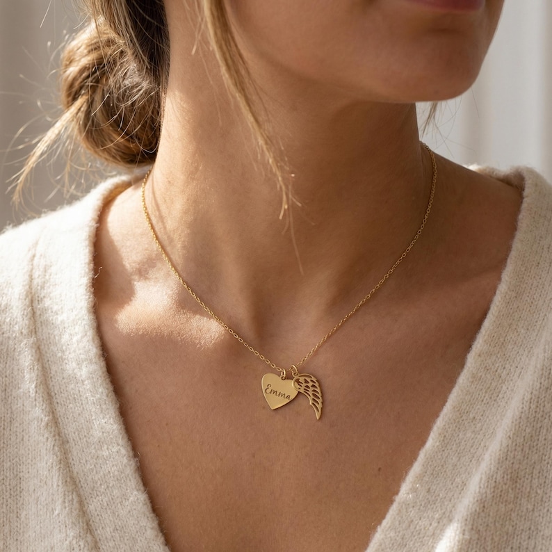 May include: A gold-colored necklace with a heart pendant and a wing charm. The heart pendant has the name "Emma" engraved on it. The necklace is worn around the neck.