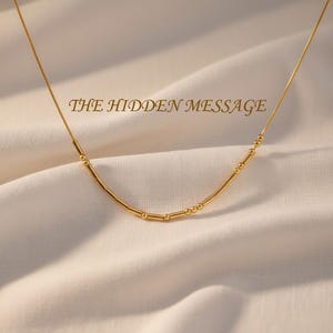 May include: A gold-toned necklace featuring a delicate chain with a central segment of alternating small round beads and short cylindrical bars arranged in a pattern, with the text THE HIDDEN MESSAGE displayed above the jewelry piece.