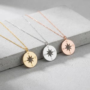 May include: Three compass pendant necklaces in gold, silver, and rose gold. Each necklace features a round pendant with a starburst design and directional markings: N, W, E, and S. The necklaces are displayed on a gray surface.