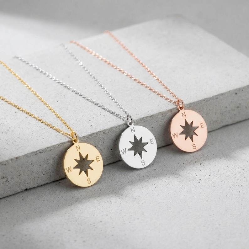 May include: Three compass pendant necklaces in gold, silver, and rose gold. Each necklace features a round pendant with a starburst design and directional markings: N, W, E, and S. The necklaces are displayed on a gray surface.