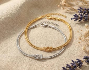 May include: Two delicate bracelets, one gold-toned and one silver-toned, are displayed. Each bracelet features a knot design at the center and a chain-like texture. The bracelets are set against a neutral background with floral accents.