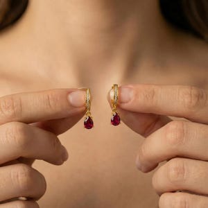 May include: A pair of gold hoop earrings featuring a small, teardrop-shaped red gemstone pendant suspended from each hoop. The hoops are accented with tiny clear stones along the front surface, creating a delicate and refined jewelry piece.