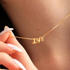 Può includere: Una collana color oro con il nome "IVY" scritto in lettere tridimensionali individuali. La collana è su una catena delicata, con le lettere ben distanziate. Lo sfondo è una vista sfocata della pelle di una persona.