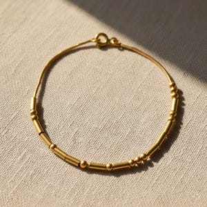 Peut inclure: Un bracelet doré délicat orné de petites perles cylindriques et rondes. Le bracelet est composé d'une fine chaîne et possède une fermeture à fermoir. Le bijou est présenté sur un tissu de couleur neutre.