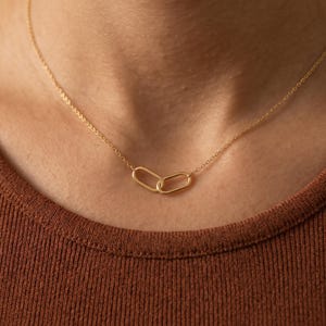 Op de afbeelding: Een goudkleurige ketting met een fijne schakelketting en twee in elkaar grijpende ovale schakels in het midden. De ketting rust op een textuurrijke, roestkleurige stof en heeft een eenvoudig ontwerp met een gepolijste metalen afwerking.
