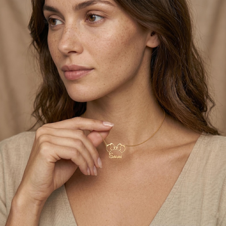 May include: A gold-colored necklace with a pendant featuring a stylized design and the name "Sarah" in a delicate script. The necklace is worn around the neck.