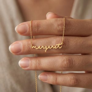 May include: A gold-toned necklace featuring a delicate chain and a central nameplate pendant displaying the name margaret in a cursive, lowercase script font. The metal has a polished finish, and the pendant is attached to the chain on both sides.