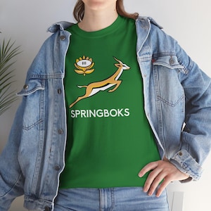 Puede incluir: Camiseta verde con el logo de los Springboks, un antílope saltando en oro y blanco, y la palabra "SPRINGBOKS" en blanco. La camiseta también tiene un emblema floral. La camiseta se usa con una chaqueta vaquera.