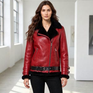 May include: A red jacket with a black faux fur collar and lining, featuring an asymmetrical silver zipper closure, two zippered side pockets, a black buckled waist belt, and buckled straps at the cuffs, worn over black pants.