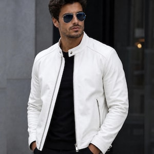 Puede incluir: Una chaqueta de cuero blanca con cuello alzado y cierre de botón a presión, cremallera frontal plateada de longitud completa y dos bolsillos laterales con cremalleras plateadas. La chaqueta se lleva sobre una camiseta negra y gafas de sol.