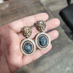 May include: Gold-toned earrings with lion head studs and red accents. The earrings have oval pendants with a dark stone centre, surrounded by a halo of small, clear stones. The earrings have a vintage design. The earrings are approximately 3 cm in length.
