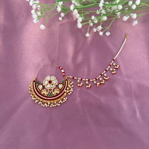 May include: A decorative gold-coloured nose ring with a crescent design. It features red, white, and gold details, a central floral motif, and dangling pearl-like beads. A delicate chain with red and white beads extends from the ring.