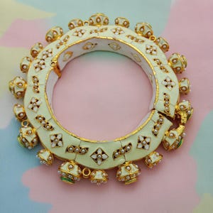 Peut inclure: Bracelet jonc articulé ivoire et doré. Le bracelet présente une base en émail crème avec des accents dorés et est orné de détails floraux et perles blancs. Il comporte des breloques décoratives dorées et crème en forme de cloche.