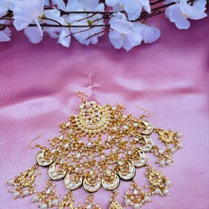 Puede incluir: Un conjunto de joyas de novia en tono dorado, que incluye una pieza para la cabeza y pendientes. La pieza para la cabeza tiene un diseño circular central con detalles de perlas colgantes. Los pendientes y la pieza para la cabeza tienen forma de media luna con detalles de perlas y oro.