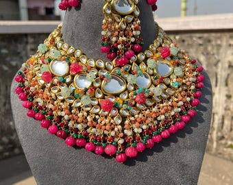 May include: A gold-coloured necklace and headpiece set featuring clear, teardrop-shaped stones, pink and green beads, and floral details. The necklace has a bib design with multiple layers of embellishments. The headpiece has a central pendant with dangling beads.
