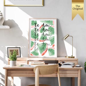 Op de afbeelding: Een ingelijste print met watermeloen schijfjes en de tekst "Te Amo". De print staat op een houten bureau met een gouden bureaulamp en een kleine potplant.