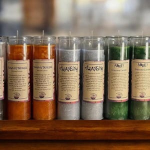 Puede incluir: Una colección de velas en tarros de vidrio con etiquetas. Las velas son de varios colores, incluyendo naranja, gris y verde. Cada vela tiene una etiqueta con texto. Las velas están dispuestas sobre una estantería de madera.