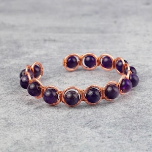 May include: Open cuff bracelet featuring deep purple amethyst beads set in a copper wire frame. The beads are evenly spaced and the copper has a warm, reddish-brown hue. The bracelet is designed to be worn on the wrist.