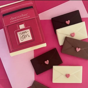 Puede incluir: Una caja de bombones belgas artesanales de Choconchoc, con el texto "Love Letters". Varios bombones con forma de sobre de chocolate negro, con leche y blanco, cada uno con un corazón rosa, están dispuestos alrededor de la caja.