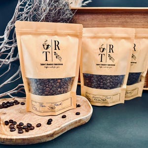 Op de afbeelding: Kraftpapieren zakken met donker gebrande koffiebonen en een transparant venster. De zakken hebben een logo met de letters "R" en "T" en de tekst "Tailor's Roastery Coffee". De zakken staan op een houten dienblad.