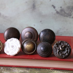May include: Assortment of ten vintage cabinet knobs in various styles and finishes, including brass, bronze, and white. The knobs are arranged on a red book.