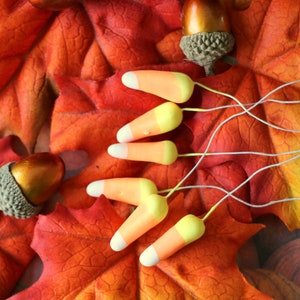 May include: Five orange and white candy corn ornaments with wire stems are arranged on a bed of fall leaves. The leaves are red, orange, and yellow.