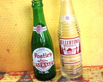 May include: Two glass bottles of soda. The bottle on the left is green and says "Postie's Delicious Beverages" with a red and white label. The bottle on the right is clear and says "Hellertown Beverages" with a red and white label.