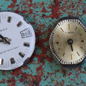May include: Two vintage Timex wristwatches with white and gold faces. The watch on the left has a white face with black hands and a date window. The watch on the right has a gold face with black hands. Both watches have black bezels.