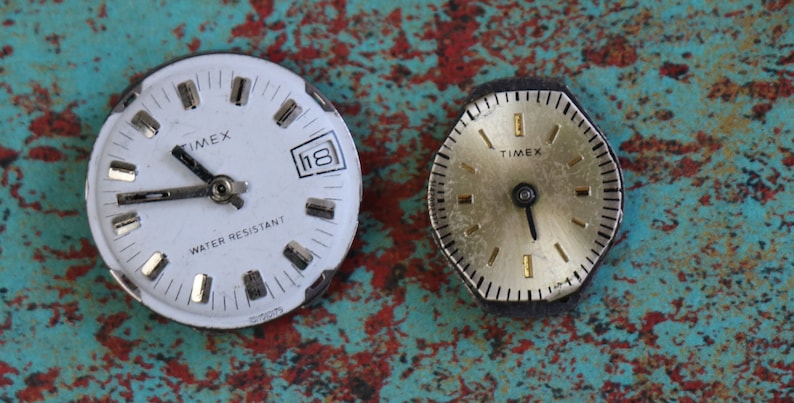 May include: Two vintage Timex wristwatches with white and gold faces. The watch on the left has a white face with black hands and a date window. The watch on the right has a gold face with black hands. Both watches have black bezels.
