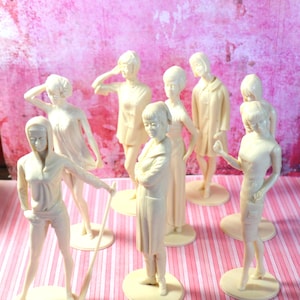 May include: A set of eight white plastic figurines of women in various poses. The figurines are standing on round bases and are arranged in a semi-circle.