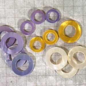 May include: A collection of iridescent, circular washers in various sizes and colors. The washers range from pale lavender to golden yellow and off-white. The washers are arranged on a light gray grid background.