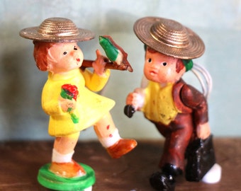 May include: Two vintage figurines of children. One is wearing a yellow dress and a straw hat, holding a bouquet of red flowers and a green branch. The other is wearing a brown suit and a straw hat, carrying a black briefcase.