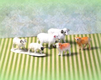 May include: A collection of miniature sheep figurines. The sheep are primarily white with black or brown markings, and one is orange. They are arranged on a green and white striped surface.