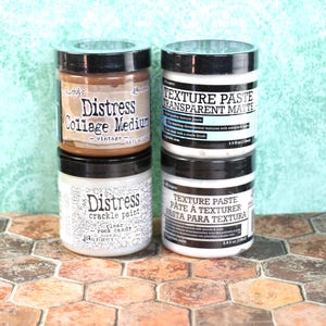 May include: Four jars of craft paint and texture paste. The jars are labeled "Distress Collage Medium - vintage", "Texture Paste Transparent Matte", "Distress Crackle Paint Clear Rock Candy", and "Texture Paste Pâte à Texturer Pasta para Textura".