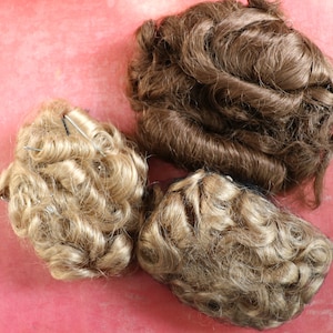 May include: Three hairpieces for doll making. One is brown and the other two are blonde. The blonde hairpieces are curly and the brown hairpiece is wavy.