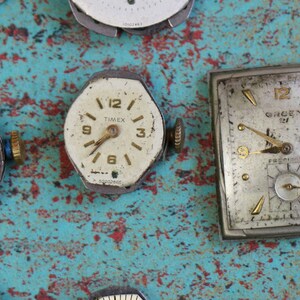 May include: A collection of vintage watch faces, some with Roman numerals, on a blue and red painted surface. The watch faces are in various shapes and sizes, including round, square, and octagonal.