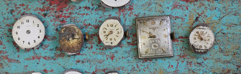 May include: A collection of vintage watch faces, some with Roman numerals, on a blue and red painted surface. The watch faces are in various shapes and sizes, including round, square, and octagonal.