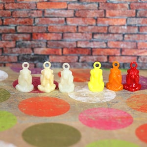 May include: Six small, colorful monkey charms in a row. The charms are in shades of off-white, yellow, orange, and red. Each charm has a loop at the top for attaching to a necklace or bracelet. The background is a brick wall and a surface with colorful polka dots.