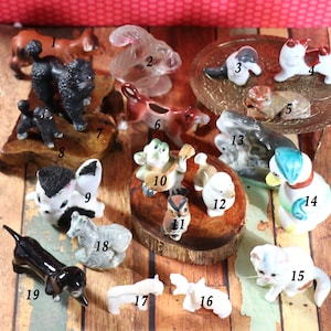 May include: A collection of 19 miniature ceramic animal figurines, including a poodle, a squirrel, a cat, a duck, a zebra, a dachshund, and an owl. The figurines are painted in various colors and are displayed on a wooden surface.