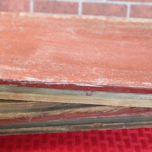 May include: Two weathered, rectangular wooden boards, one stacked atop the other. The top board is painted a faded red, showing signs of wear. The bottom board has a mix of natural wood tones. The boards rest on a red surface.