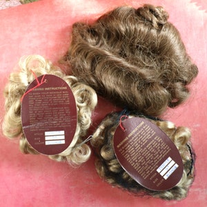May include: Three doll wigs with brown and blonde hair. Each wig is attached to a brown card with the text "Washing Instructions" and the words "Stock", "Size", and "Color" with the corresponding information filled in. The wigs are on a pink surface.