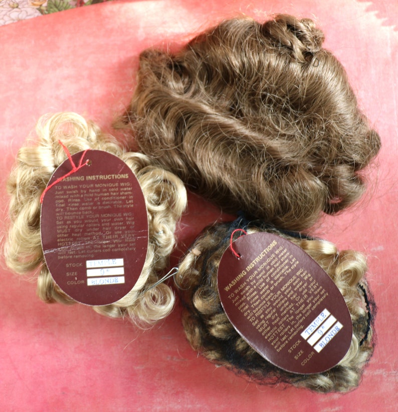 May include: Three doll wigs with brown and blonde hair. Each wig is attached to a brown card with the text "Washing Instructions" and the words "Stock", "Size", and "Color" with the corresponding information filled in. The wigs are on a pink surface.