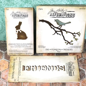 May include: Three die-cutting templates for crafting. The first shows a chocolate bunny with a white bow. The second shows a bird on a branch. The third shows the word "FRIENDS" in a stylized font. All three templates are from the Jim Holtz Alterations Movers & Shapers collection.