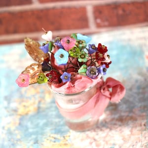 May include: A small glass vase filled with a colorful bouquet of glass flowers. The flowers are in shades of pink, blue, green, and red. The vase is wrapped in a pink ribbon.