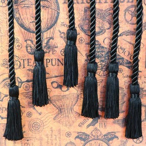 May include: Black tassels with twisted cords, displayed against a steampunk-themed background. The tassels are made of a silky material and are attached to a thick, twisted cord. The background features vintage illustrations and text.