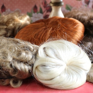 May include: Assortment of doll hair pieces in various colors and textures, including blonde, brown, and red. The hair pieces are arranged on a pink and white patterned surface.
