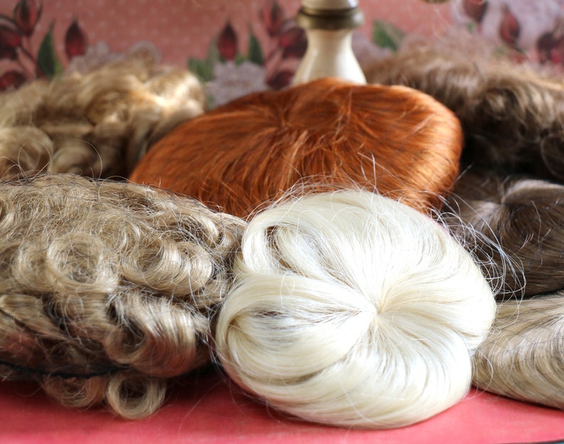 May include: Assortment of doll hair pieces in various colors and textures, including blonde, brown, and red. The hair pieces are arranged on a pink and white patterned surface.