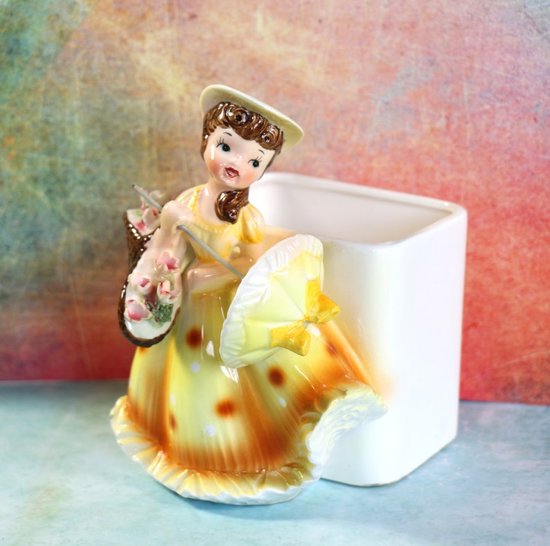 May include: A vintage ceramic planter shaped like a girl in a yellow dress with white polka dots, holding a yellow umbrella and a basket of flowers. The planter is white and has a square shape.