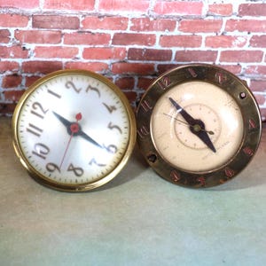 May include: Two vintage clocks with gold-colored metal casings. One clock has a white face with black numerals and hands, and a red second hand. The other clock has a cream-colored face with black hands and numerals.
